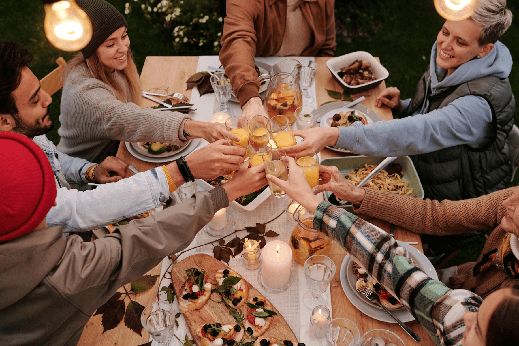 A group of friends sitting at an outdoor dinner table set with food are raising their glasses together to cheers.