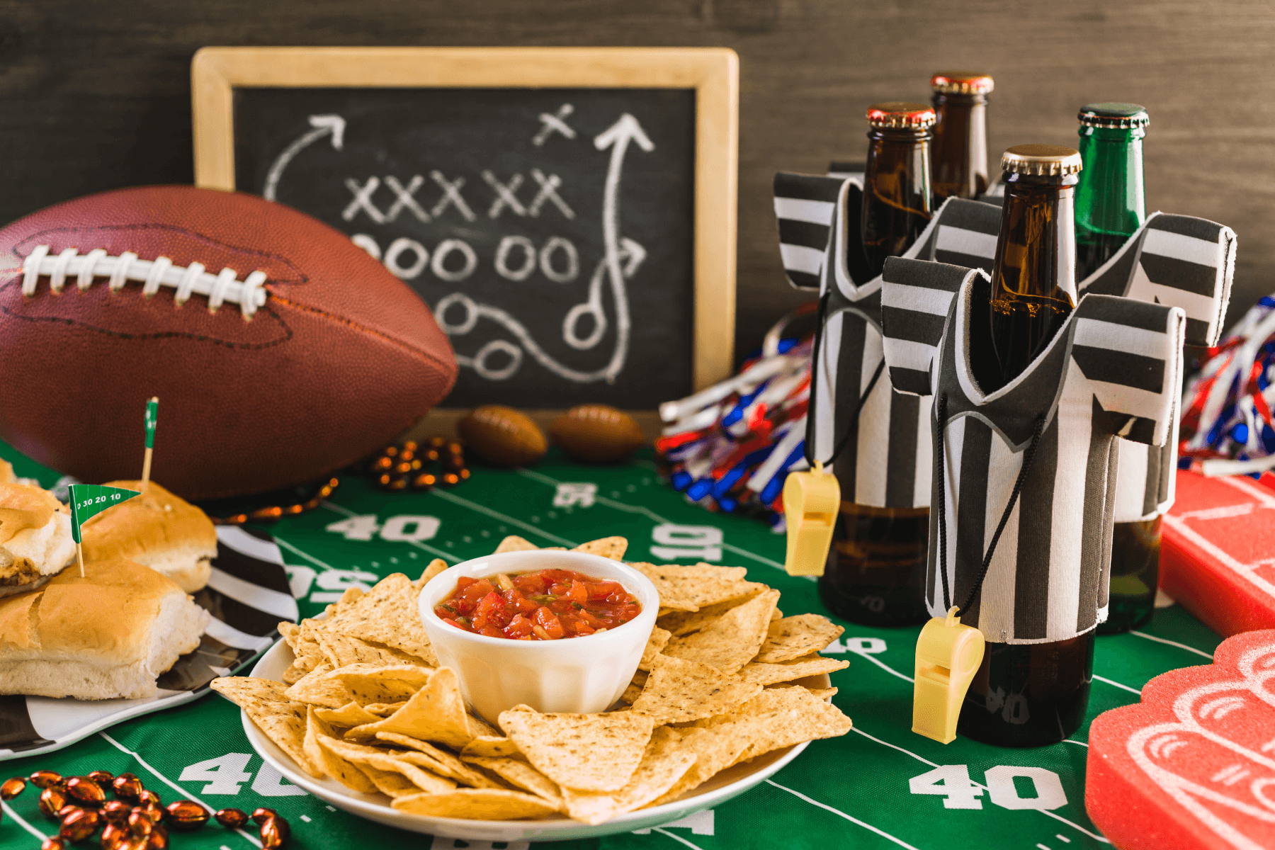 A table set with chips and salsa, bottles of beer with referee-style koozies and whistles, a plate of sliders, a football, a miniature chalkboard, and a green tablecloth resembling a football field.