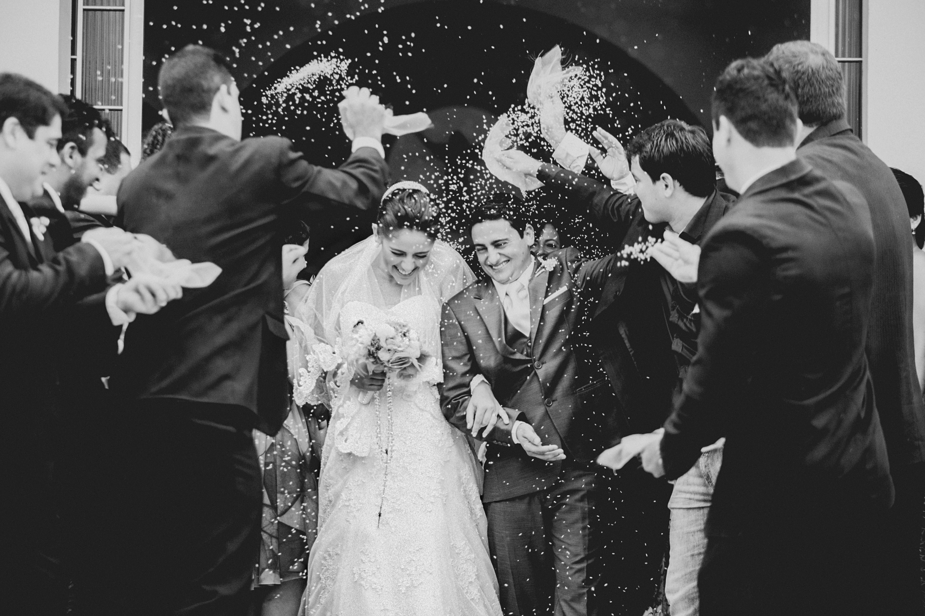 A black and white photo of a bride and groom being showered in confetti as they walk out of their wedding together.