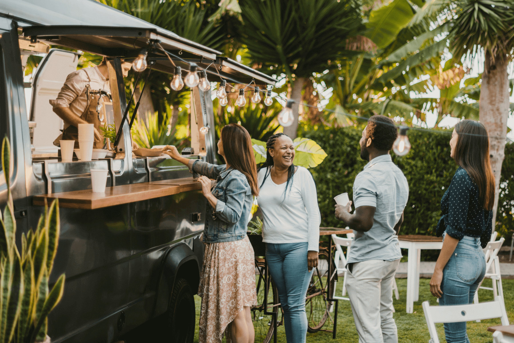 A line of people standing at a food truck in a yard set with tables and chairs.