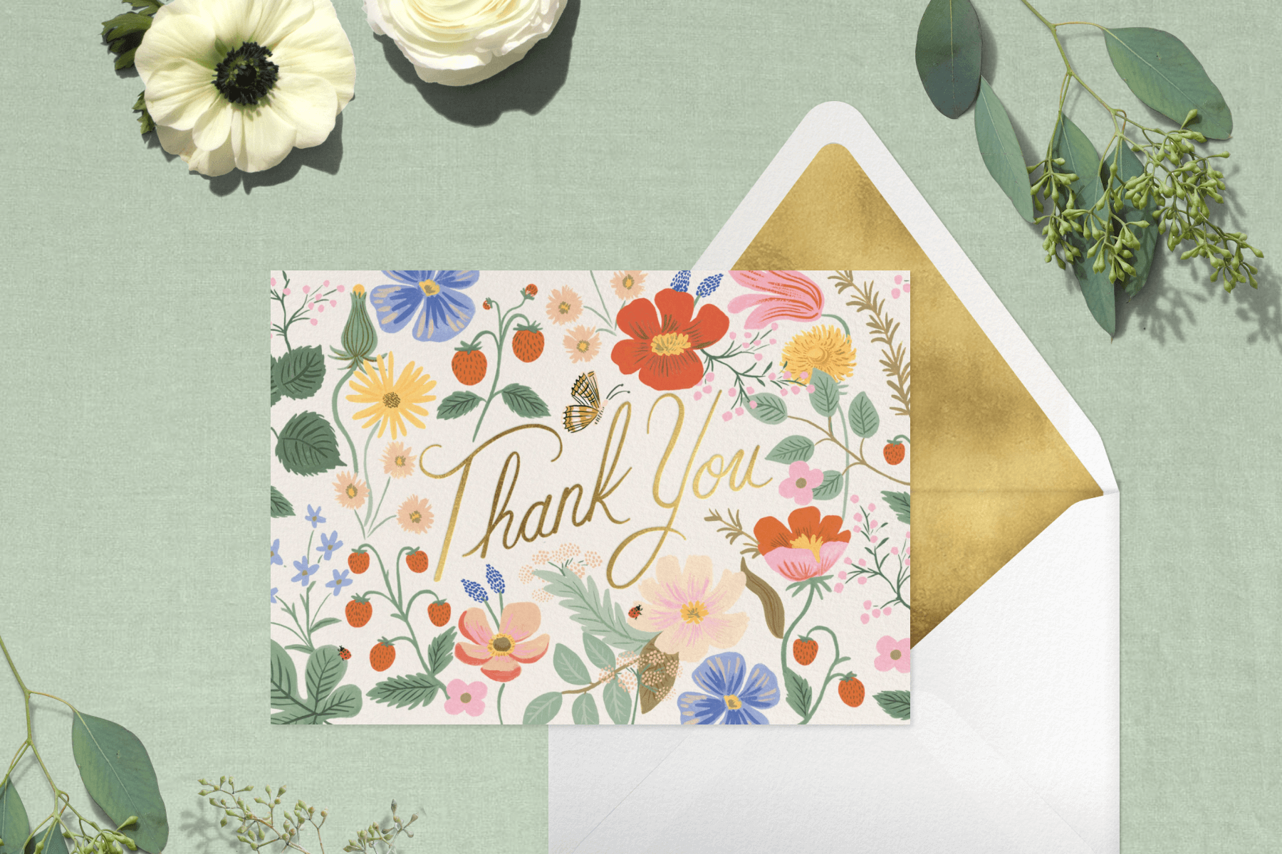 A thank you card with illustrated wildflowers and strawberries and ‘thank you’ in gold script with a matching envelope on a sage green backdrop with white flowers and greenery.