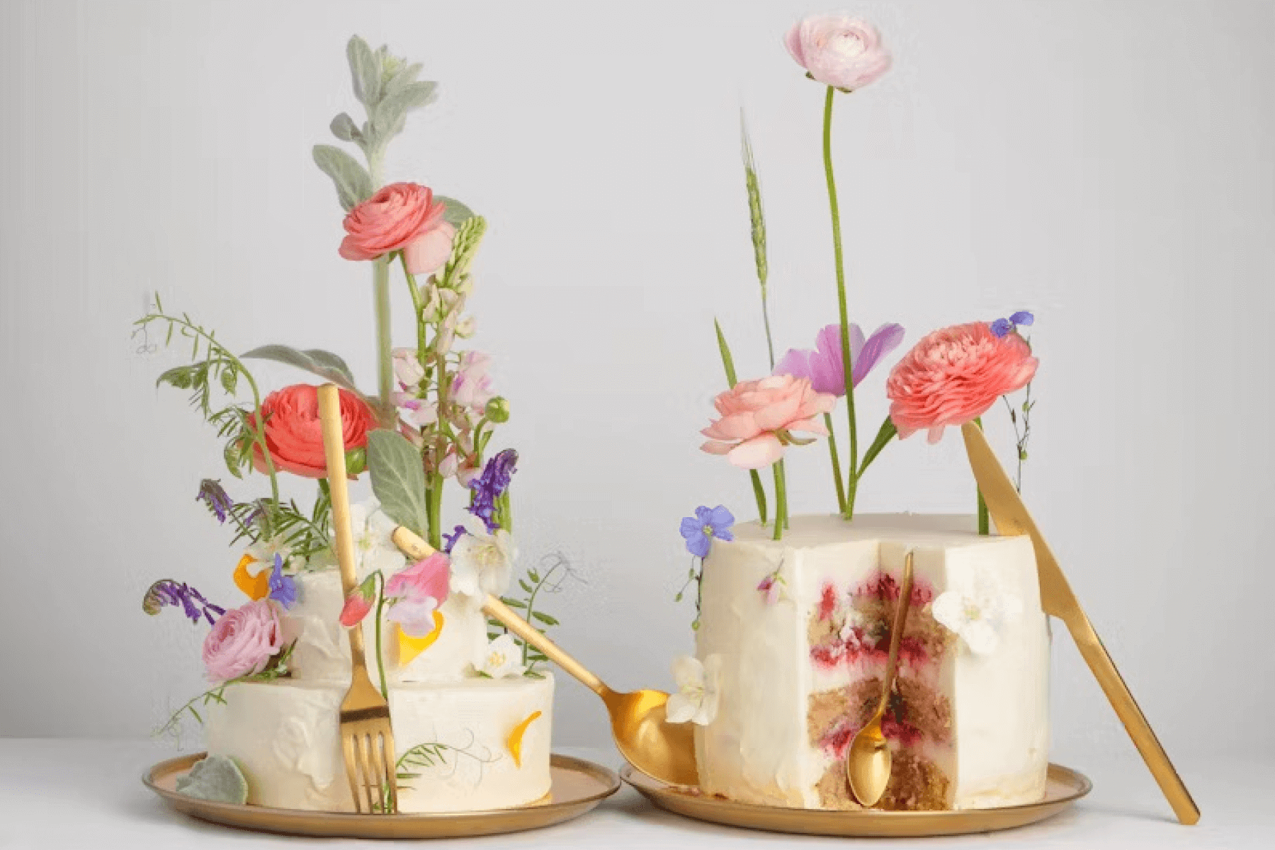 Two wedding cakes covered in colorful flowers on gold plates with gold silverware.