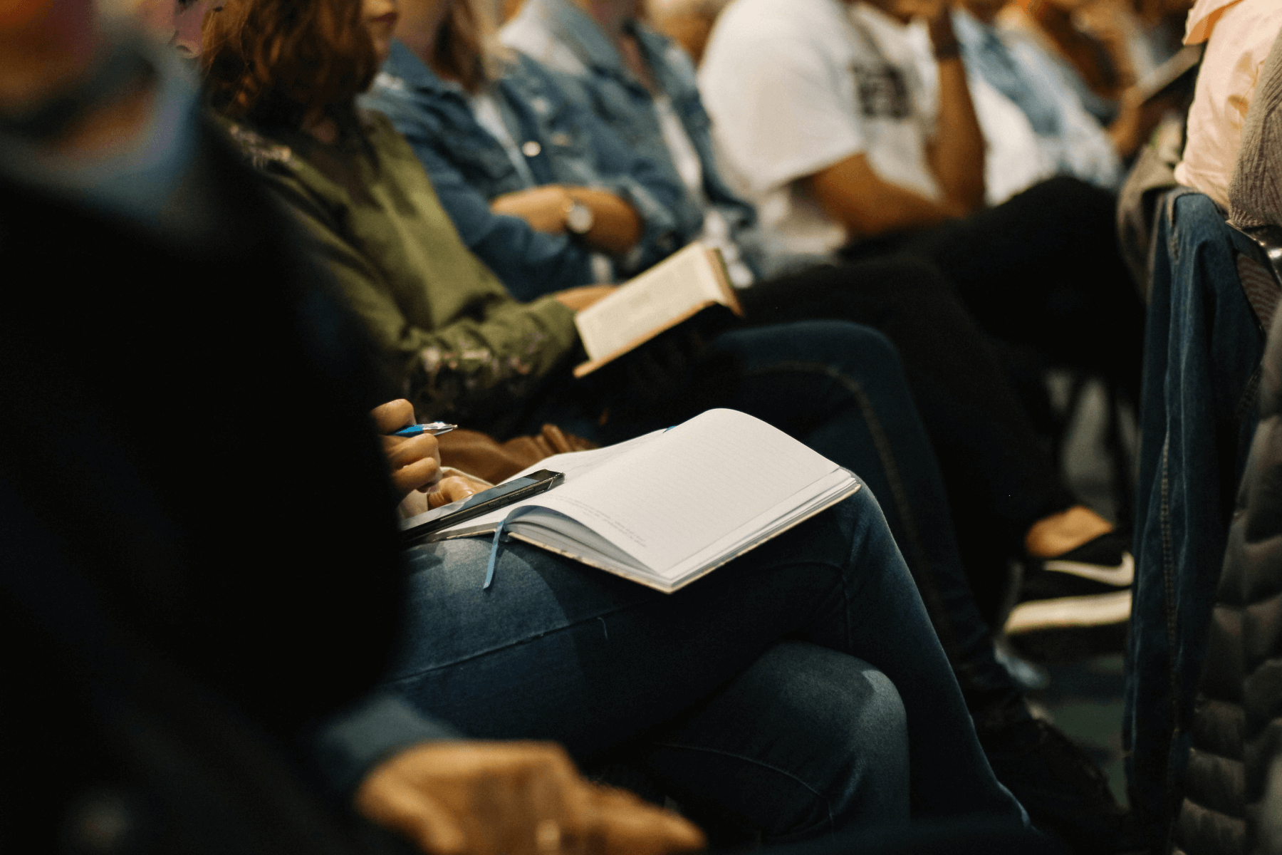 An audience sitting in chairs at an event, holding notebooks, and taking notes.