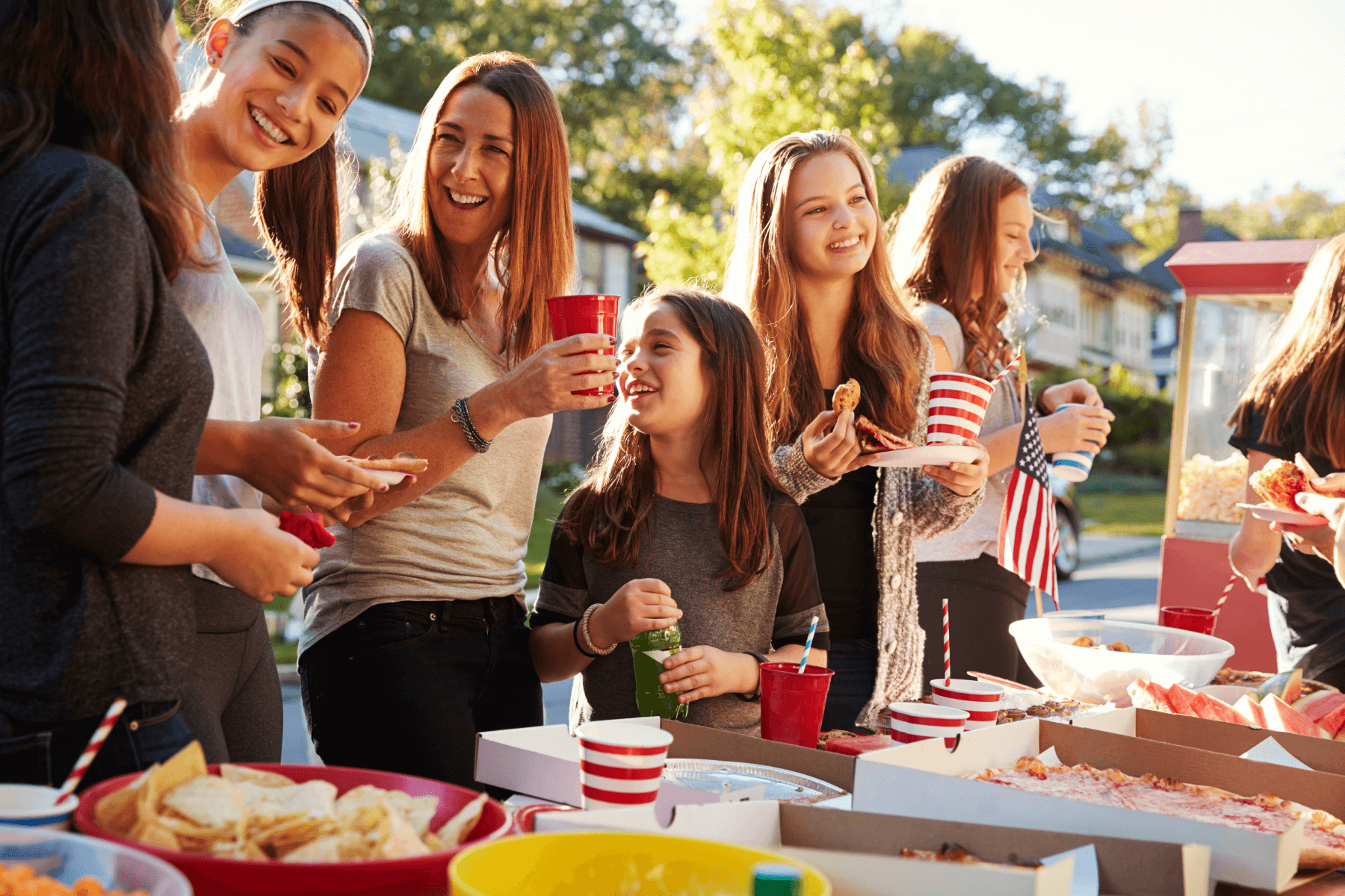 Several women laugh and talk in front of a table of food.