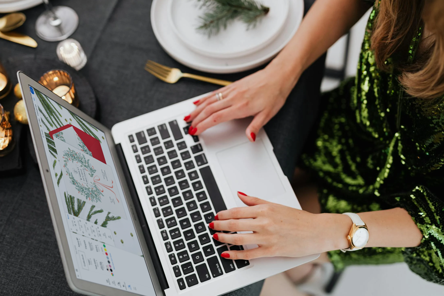 A woman wearing a sequined green dress and red nail polish designs a Paperless Post Christmas party invitation on a silver laptop.