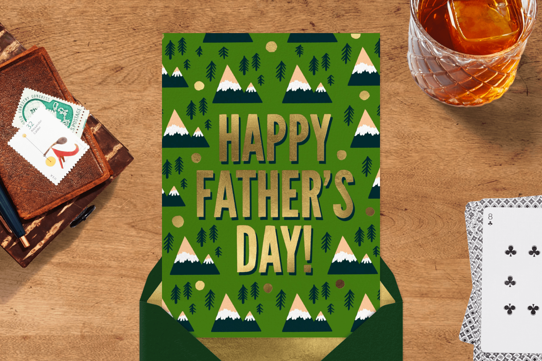 A green card reads HAPPY FATHER’S DAY! in gold letters surrounded by simple mountains and trees, on a wooden backdrop with stamps, an adult beverage, and playing cards.