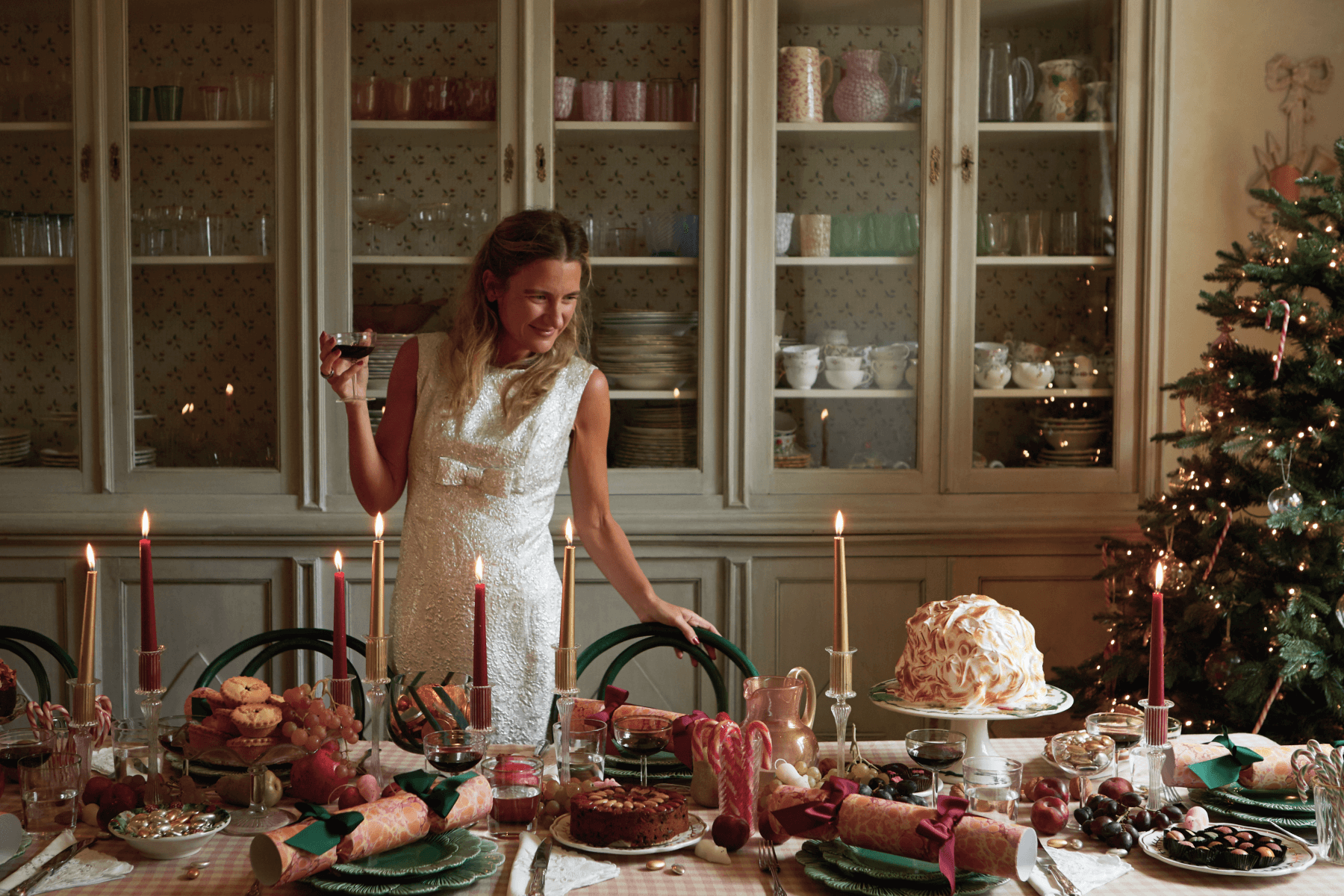 Skye McAlpine holding a cocktail while standing over an elaborately set dining table with candles, holiday décor, cakes, fruit, and candy.