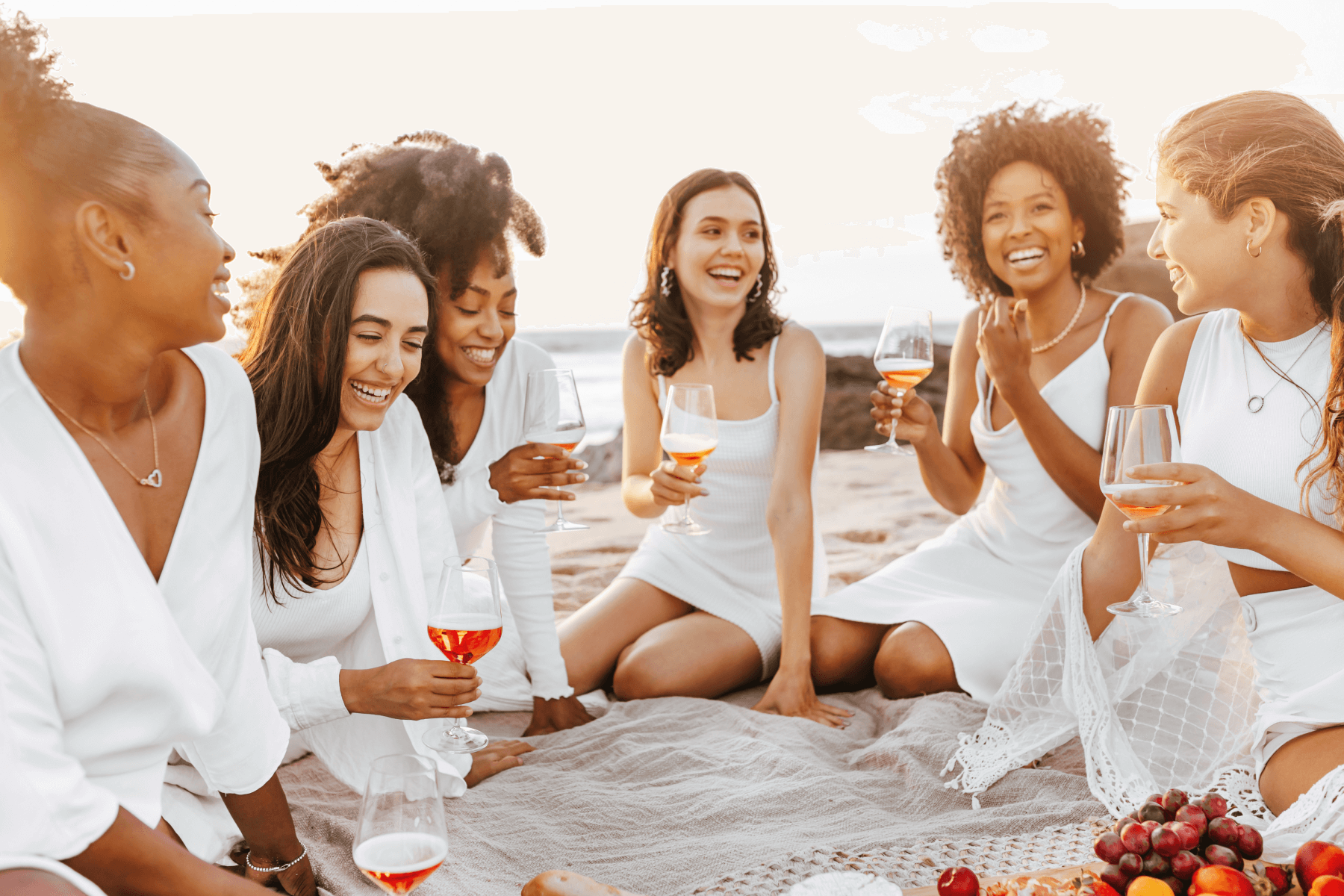 A group of women sit on a beach, wearing white and drinking wine.