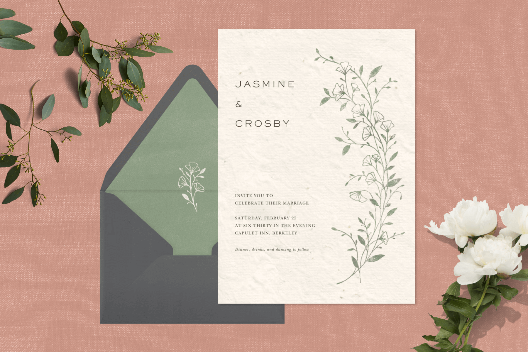 An off-white wedding invitation with a branch of greenery and a matching envelope with a green liner over a beige backdrop with white flowers and green leaves.