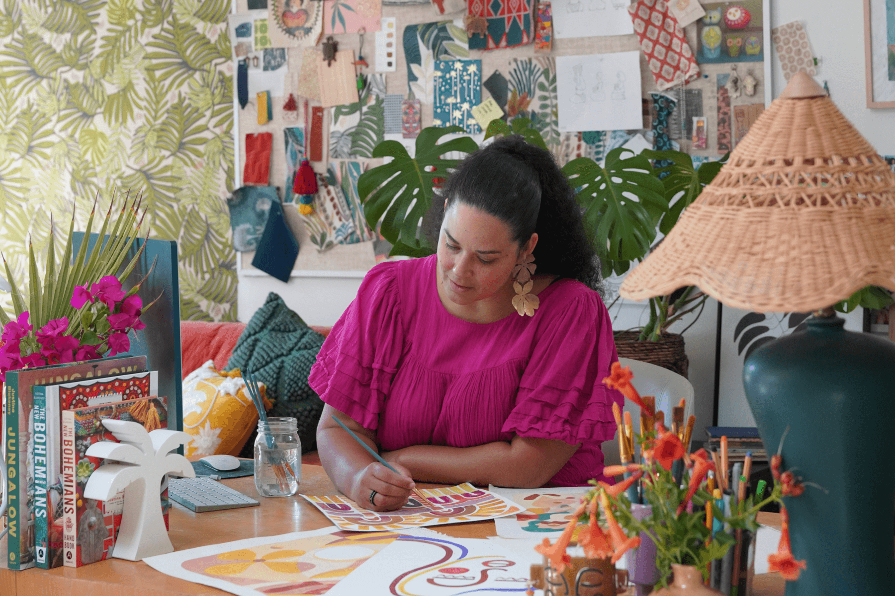 Jungalow founder Justina Blakeney sits at her desk, painting colorful designs, surrounded by plants and colorful decorations.