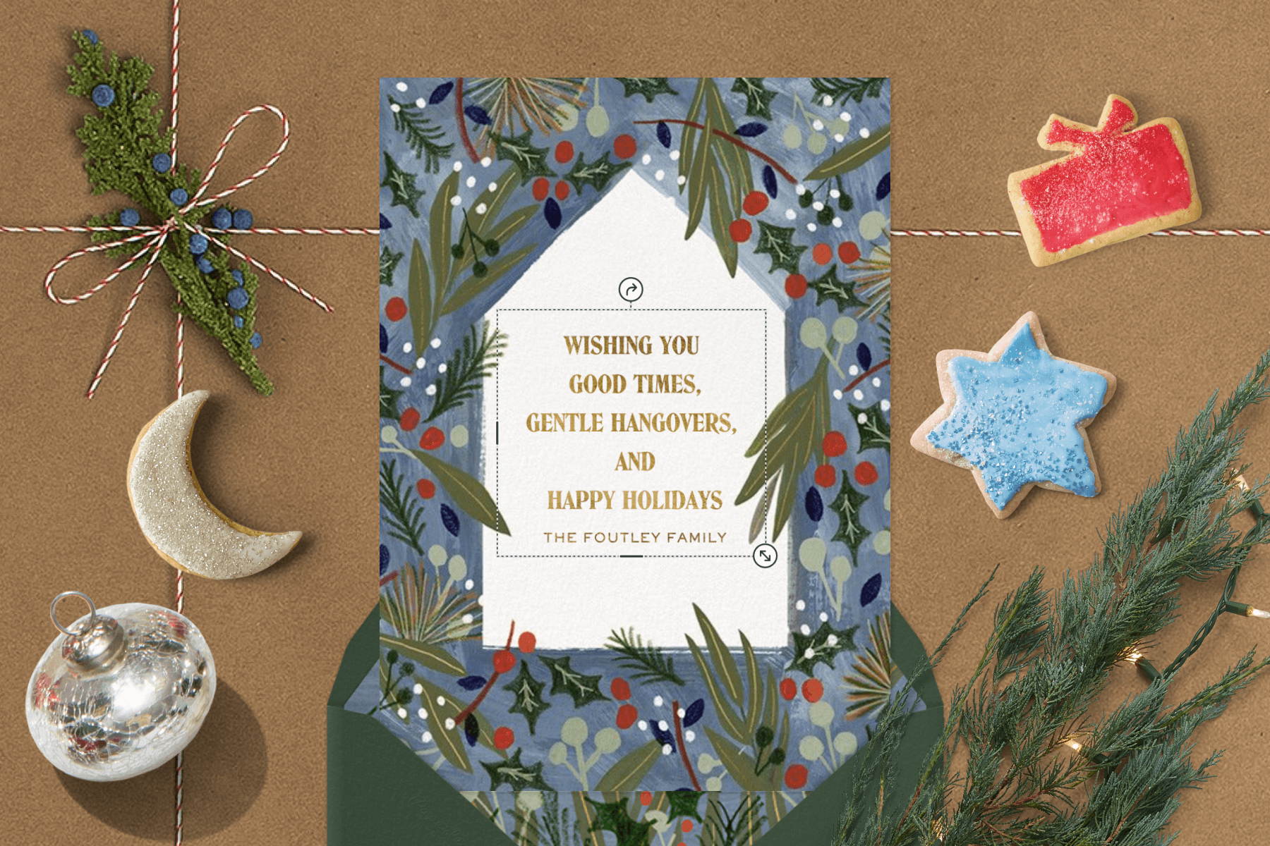 A card with a simple house shape and holly berries on a blue background, next to Christmas cookies, string lights, and decorative string.