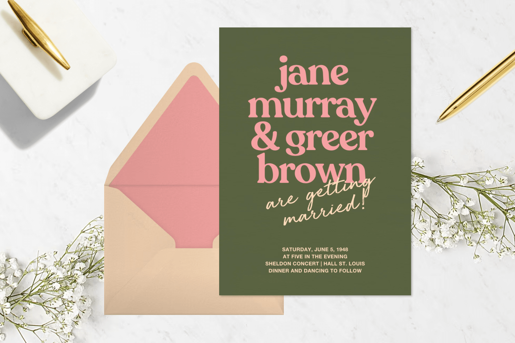 A dark green wedding invitation with the couples’ names in pink text in front of a peach envelope, over a marble background decorated with white flowers and a gold pen.