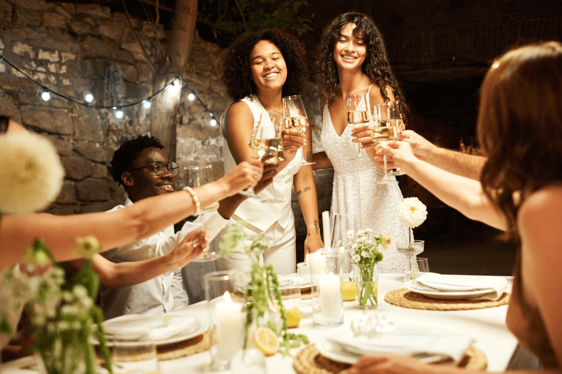 Two women wearing white stand and cheers glasses of wine with friends over an outdoor table set with white flowers surrounded by string lights.