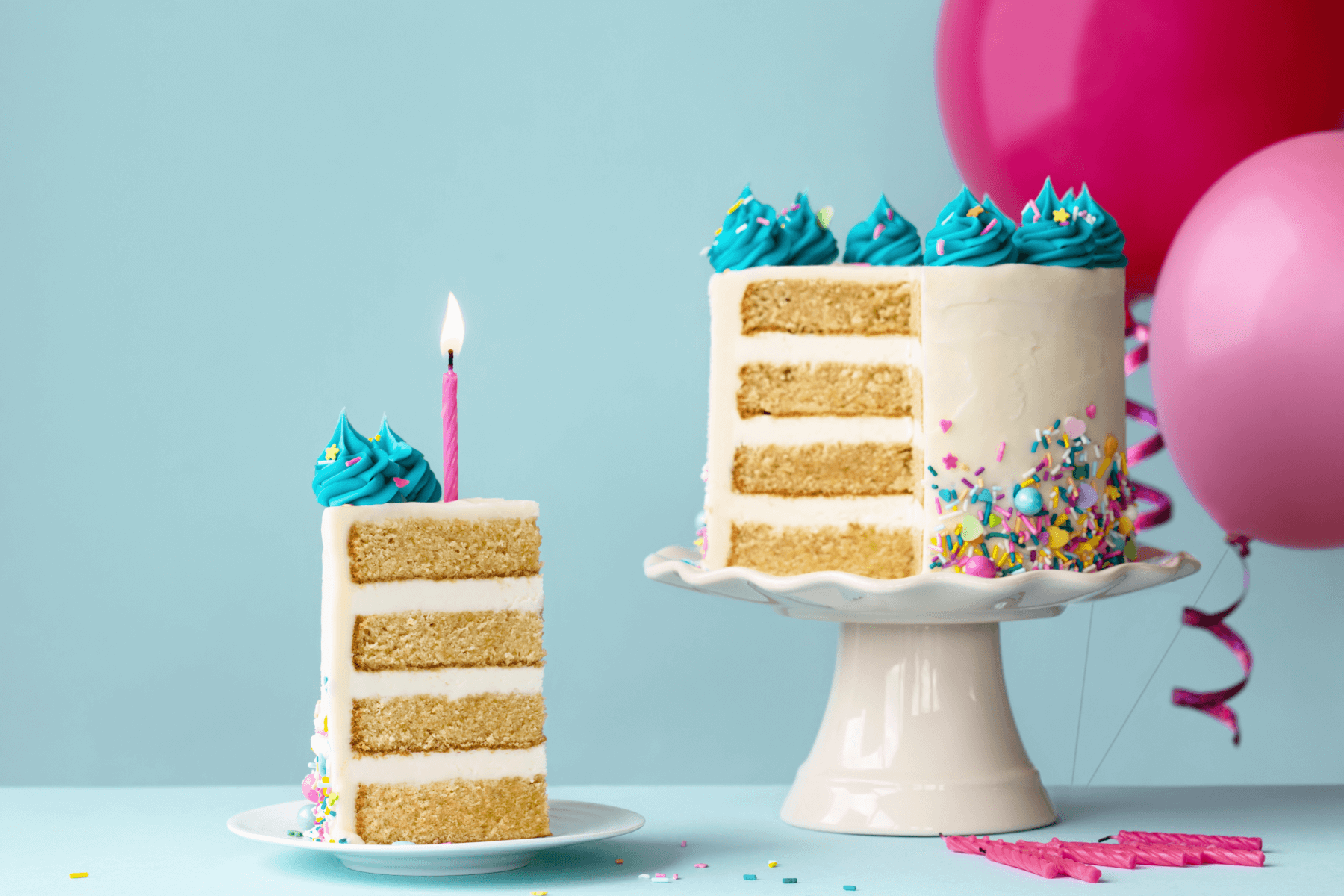 A slice of cake sits on a teal table with a teal background; next to it, the rest of the cake sits on a white ceramic tray with pink balloons in the background.