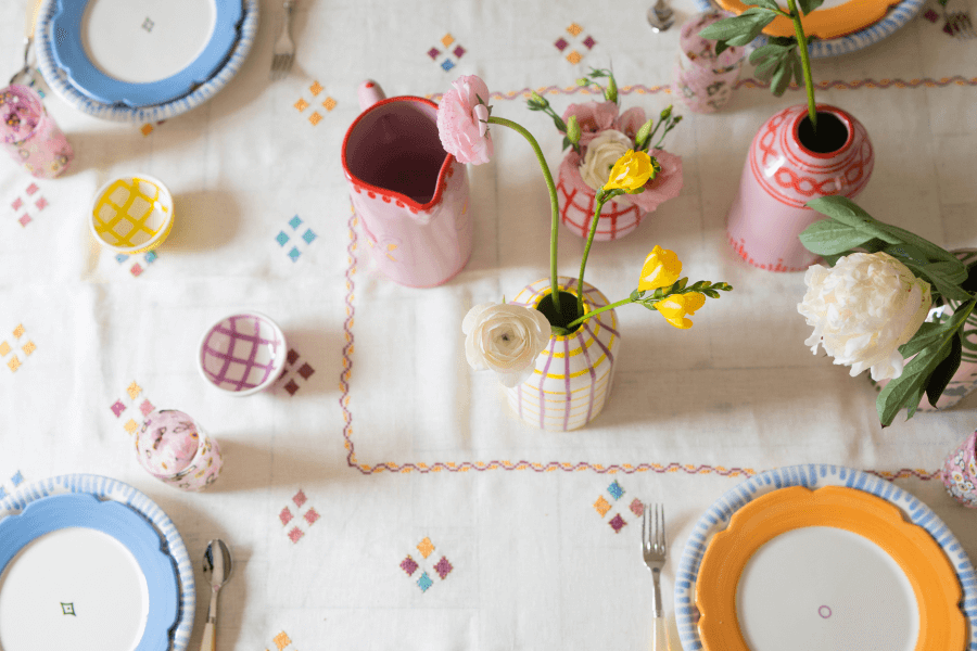 A table set with white, blue, and yellow plates, pink vases with flowers, and a white tablecloth with a colorful diamond pattern.