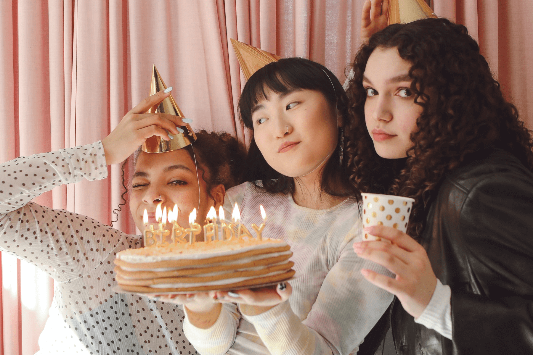 Three girls wear golden birthday hats and pose with a birthday cake made out of pancakes and a sparkly polka dot cup in front of a pink curtain.