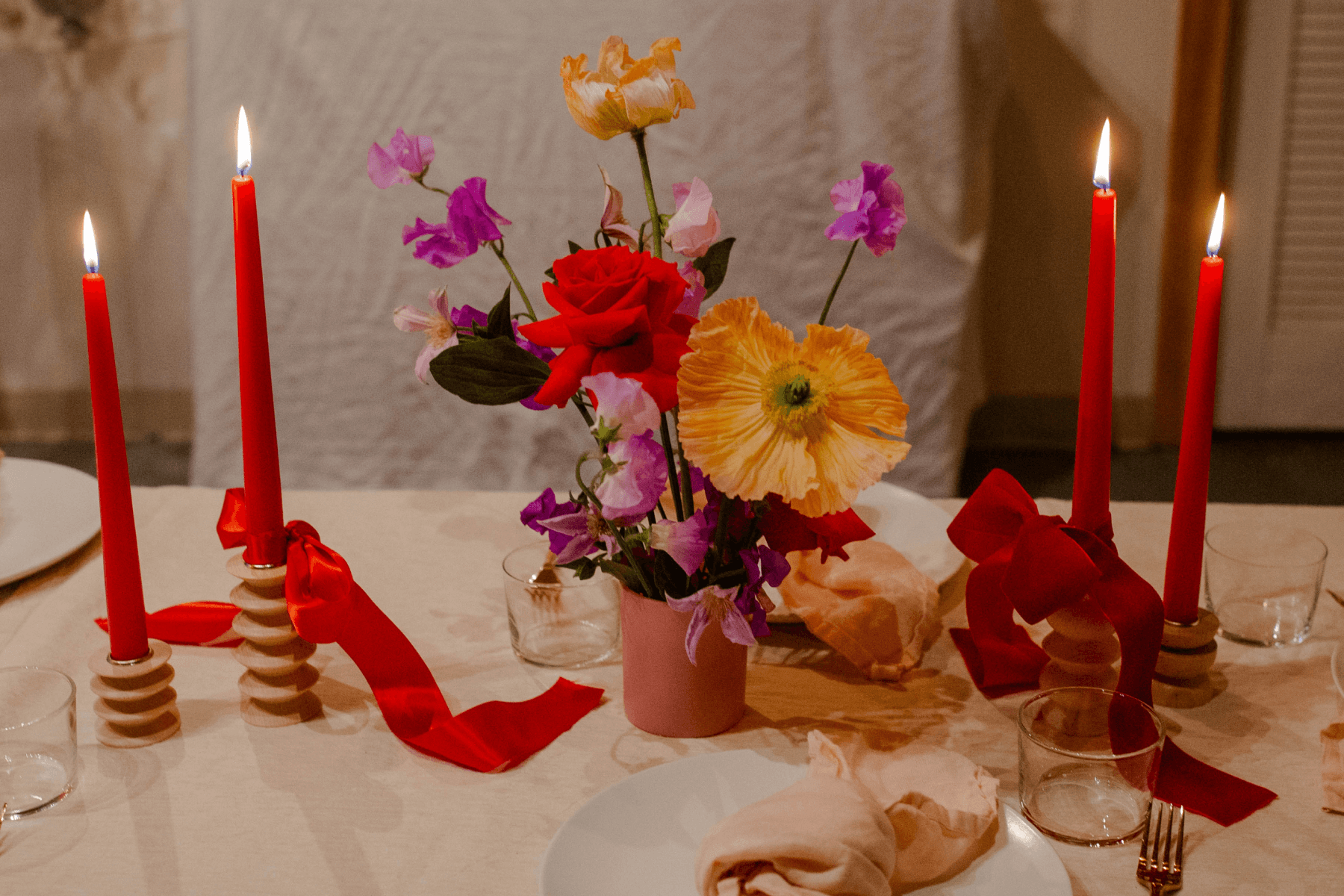 A table set with red candles with red ribbons and a centerpiece of red, yellow, and purple flowers.