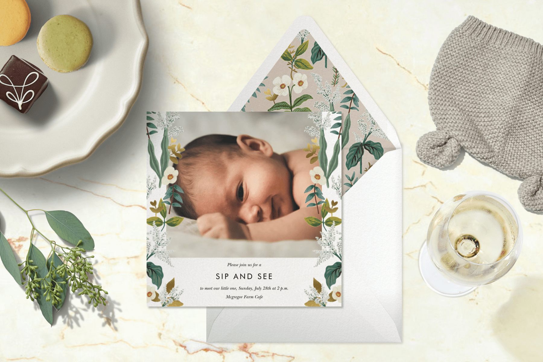 A sip and see invitation featuring a floral border and a photo of the new baby over a marbled background set with greenery, a glass of white wine, a knit baby beanie, and a plate of various desserts.