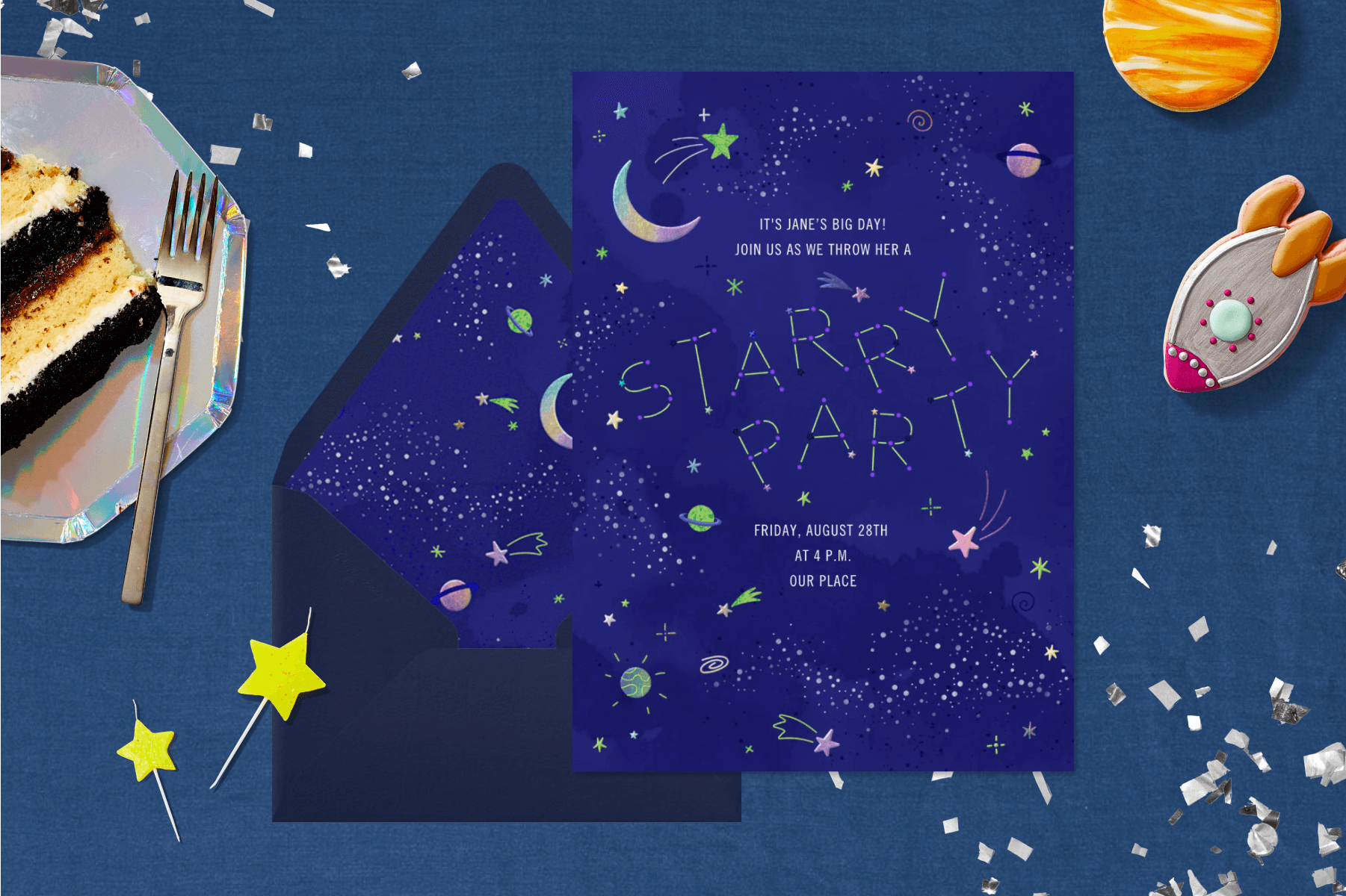 A dark blue birthday invitation with stars and planets and an envelope with a matching liner with stars and planets, all over a blue background with space-themed cookies, a slice of cake, star-shaped candles, and silver confetti.