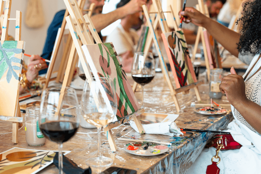 People painting at individual easels set on a table with wine glasses.