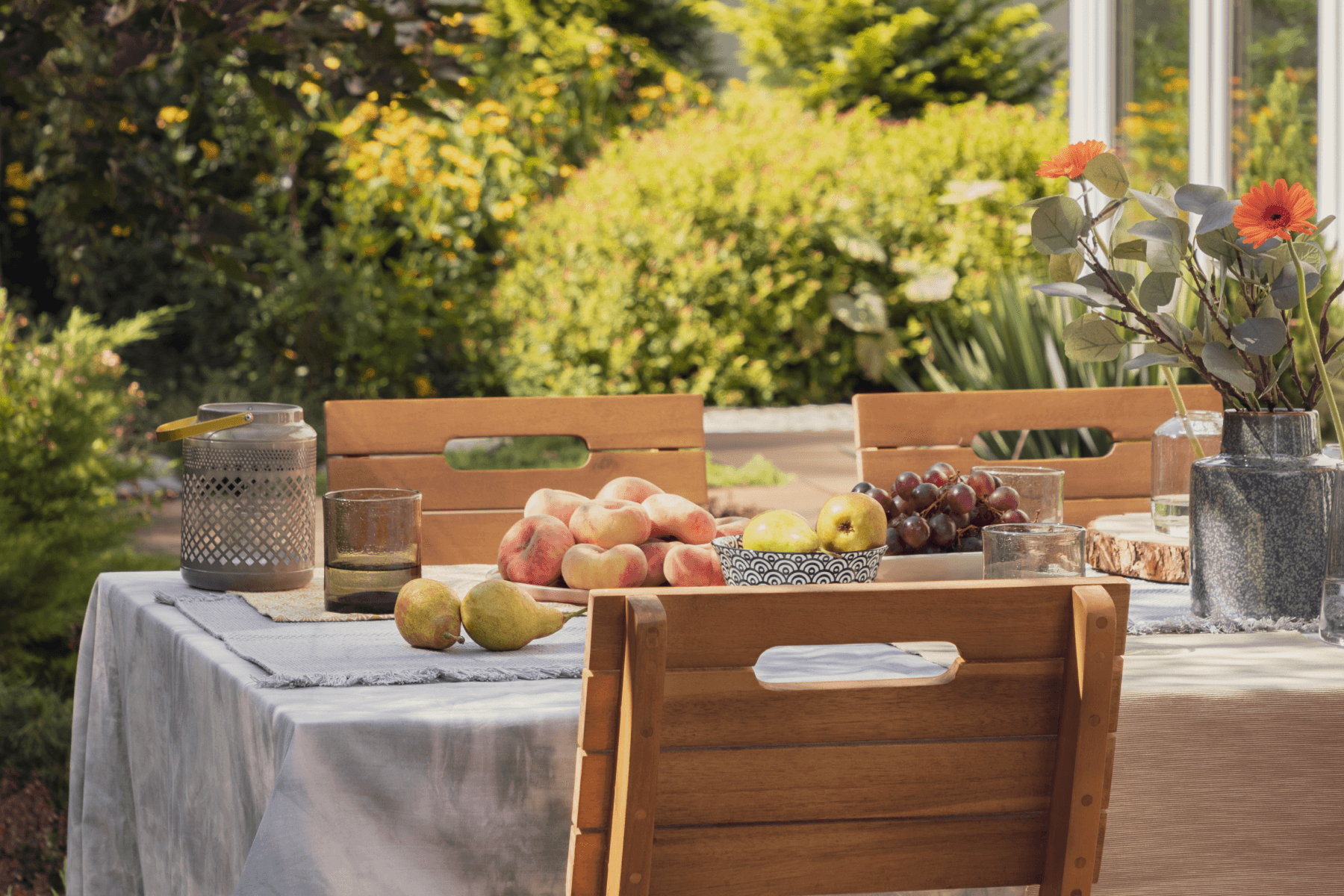 Wooden chairs around an outdoor table set with bowls of various fruits and vases of flowers