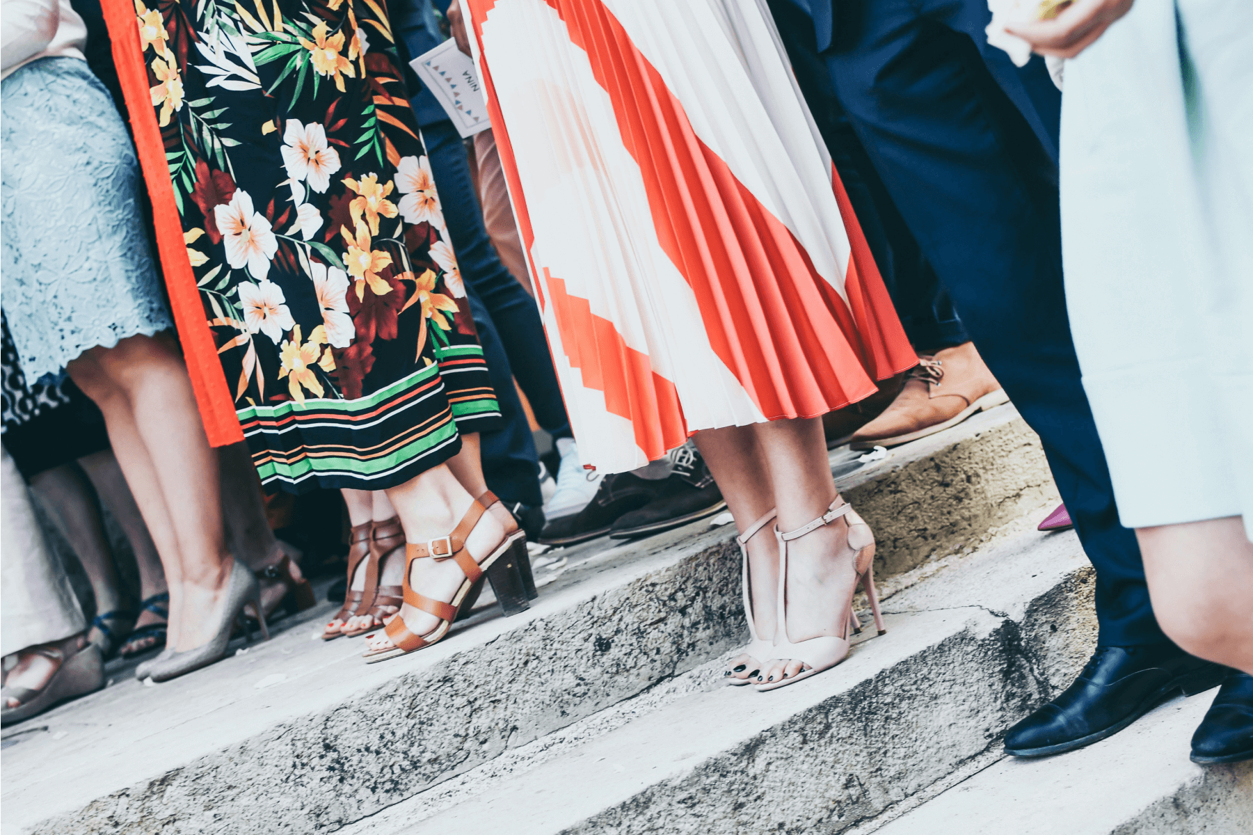 A close-up of several pairs of legs standing on steps at an event wearing dresses, high heels, suits, and dress shoes.