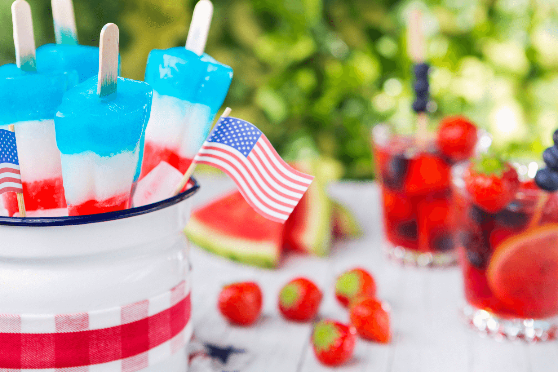 Red, white, and blue popsicles in a bucket with an American flag and strawberries, watermelon, and red fruity cocktails in the background.