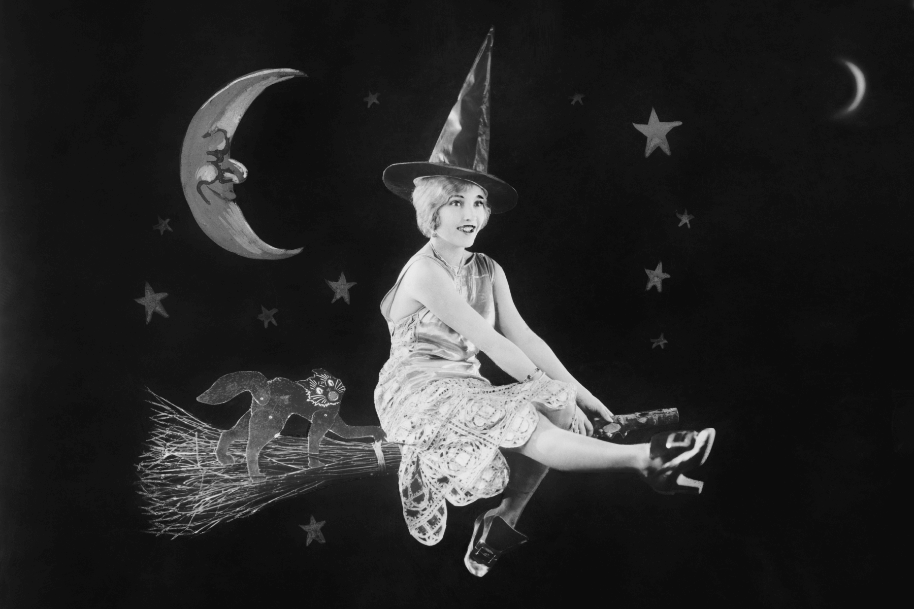 A model dressed as a witch rides a broom in front of a fake night sky.