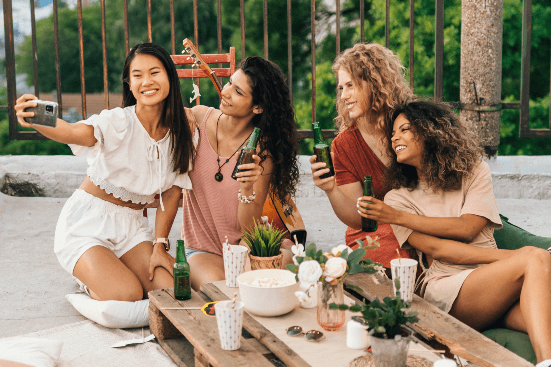 A group of women sit around a picnic table and take a selfie while holding beer bottles.