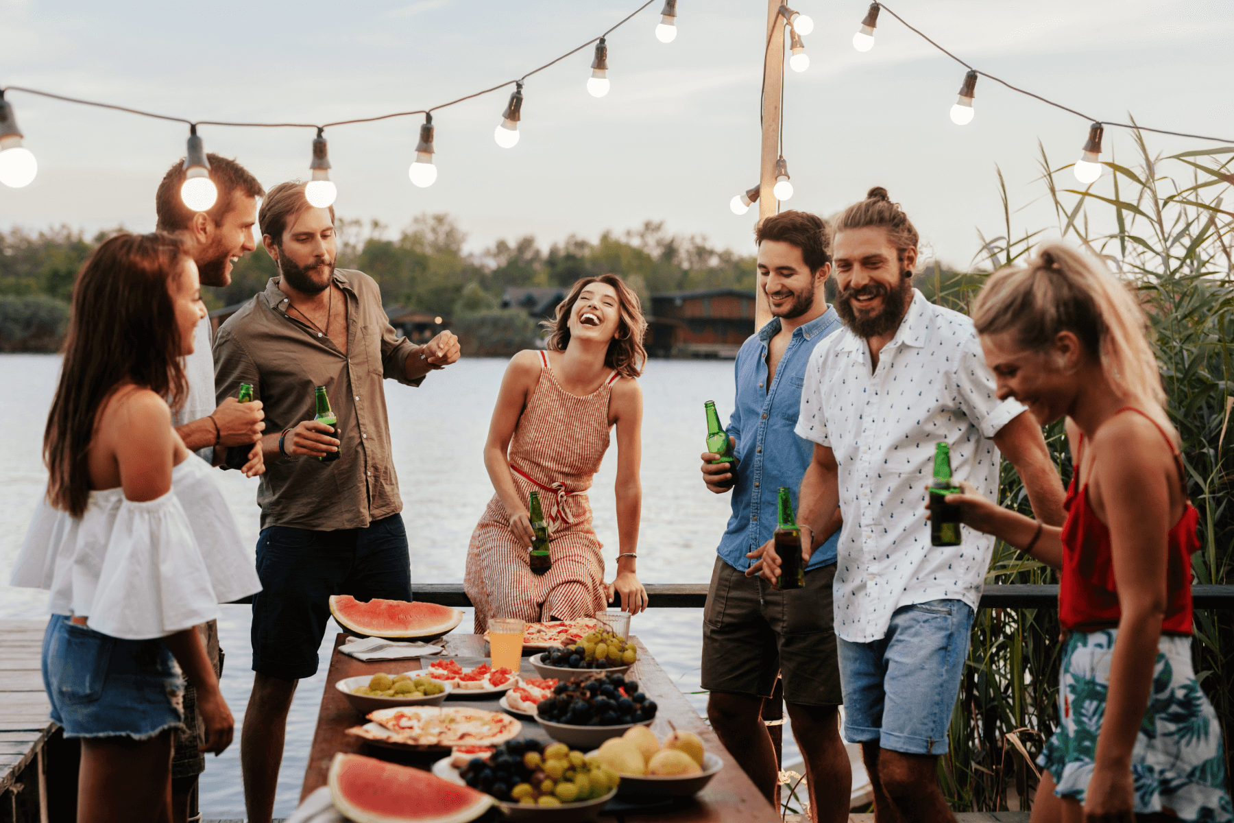 A group of friends by a lake, laughing and drinking beers around a table of watermelon and other party snacks.