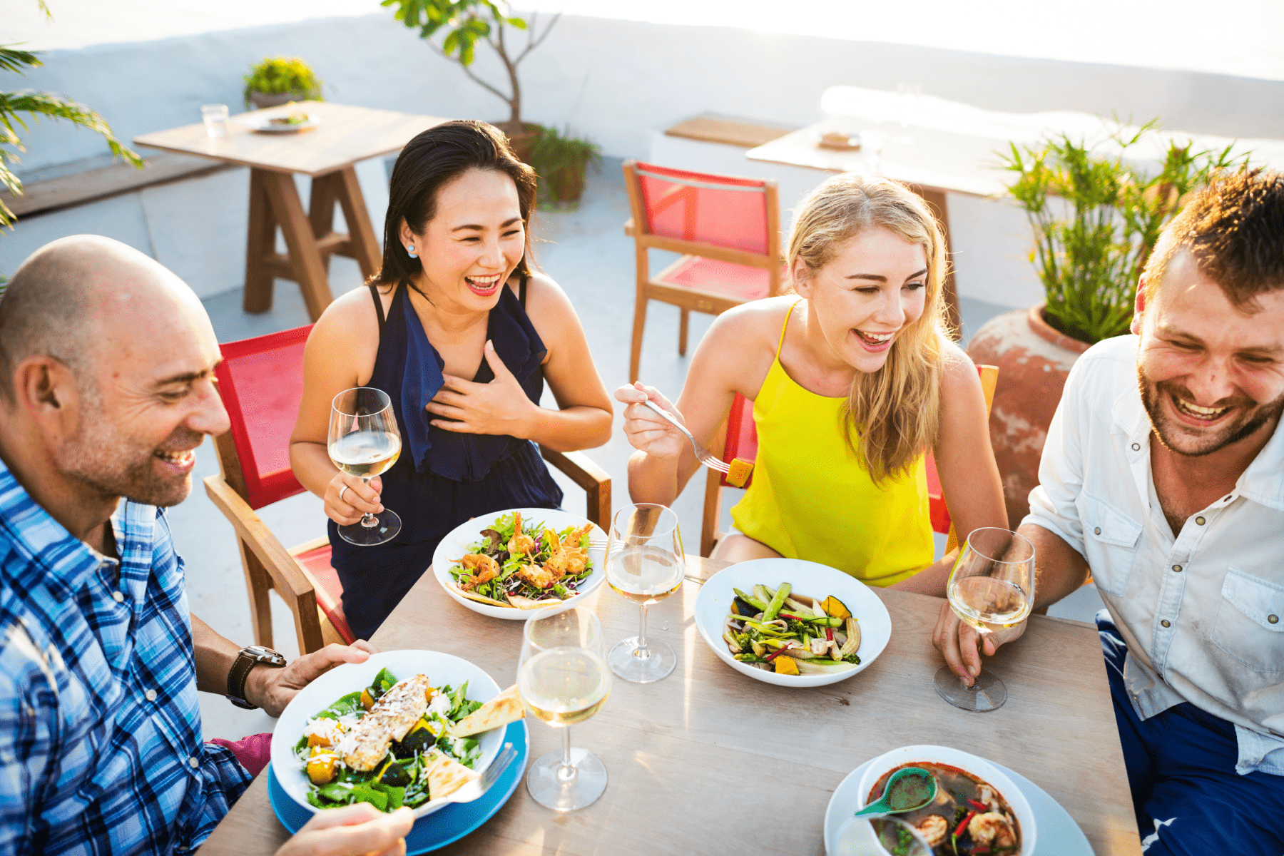 Four friends laughing together at a table with plates of food and glasses of wine.
