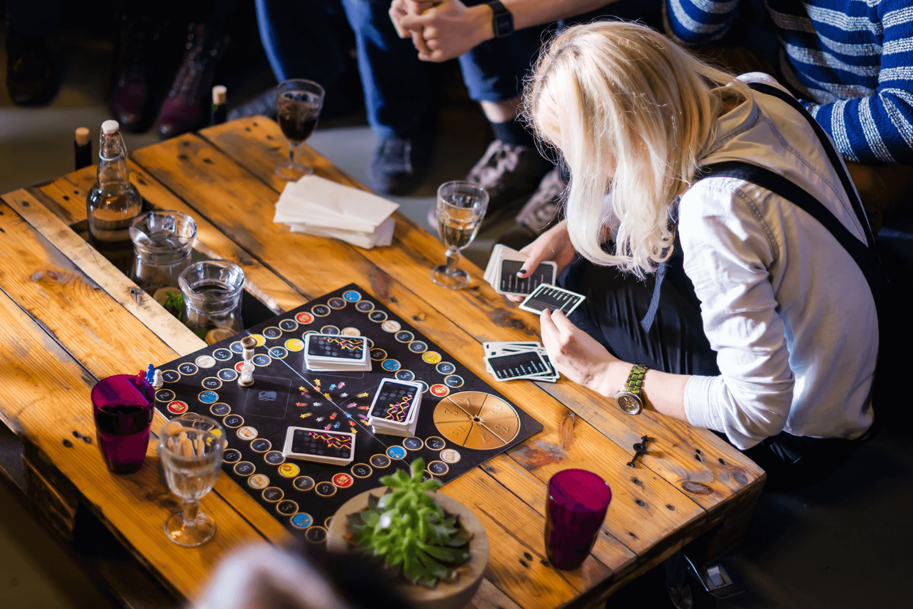 Friends sitting around a wooden table with cups and wine glasses while playing a board game. 