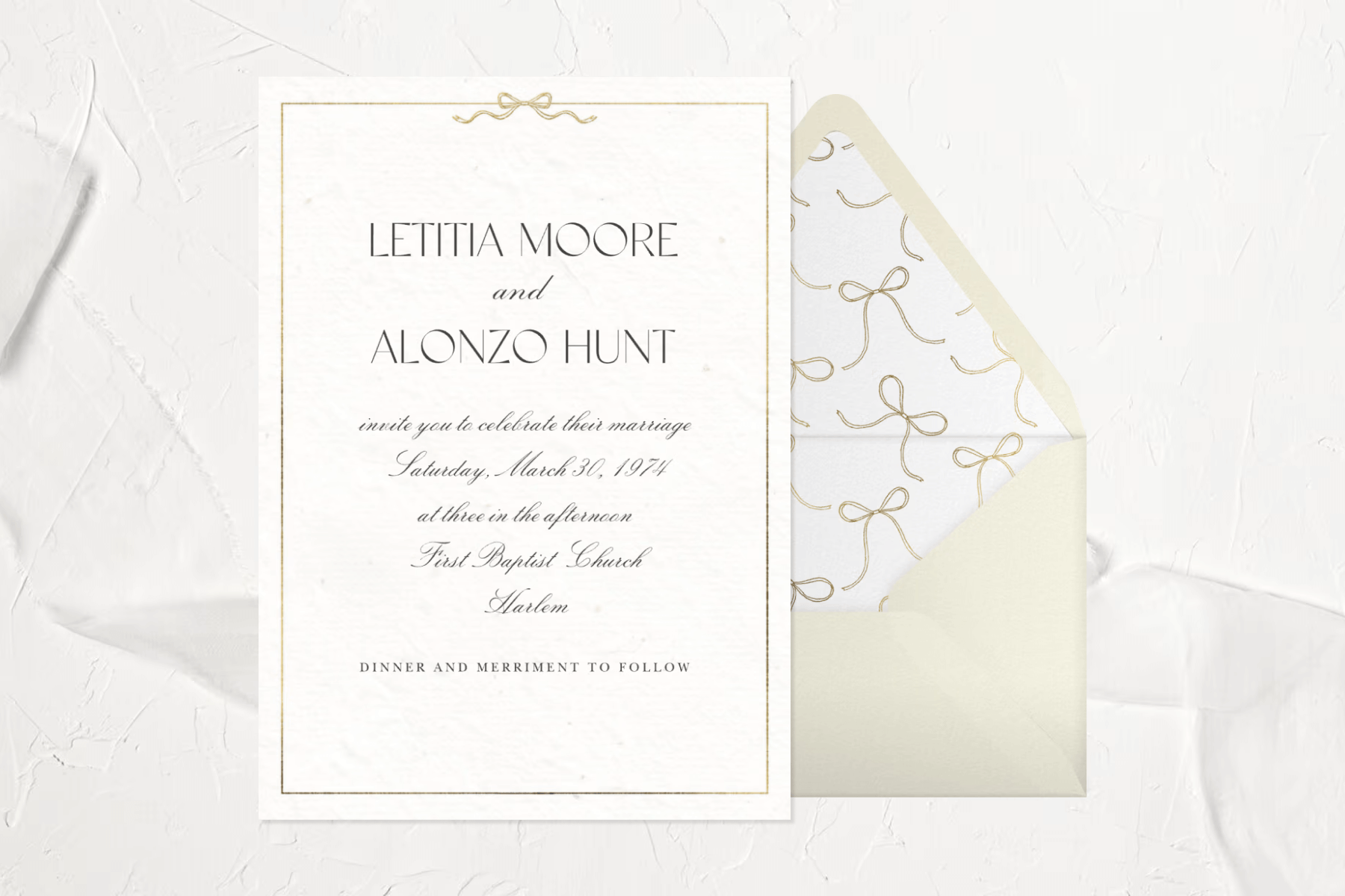 A white wedding invitation with a gold border featuring a small bow at the top in front of an envelope with a bow-themed liner. 