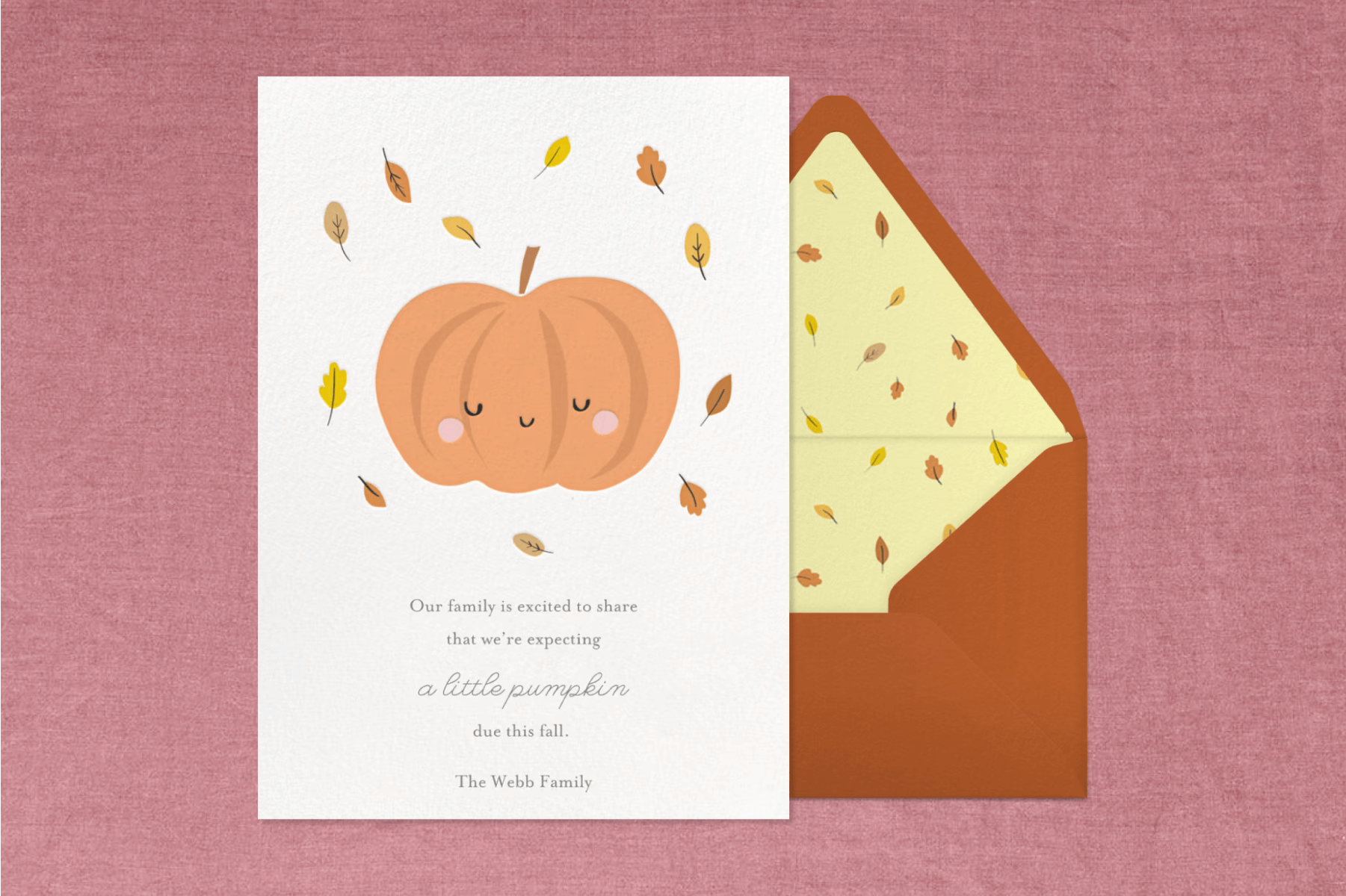 A pregnancy announcement card with an illustration of a smiling pumpkin and falling leaves next to a matching orange envelope and in front of a burgundy backdrop.