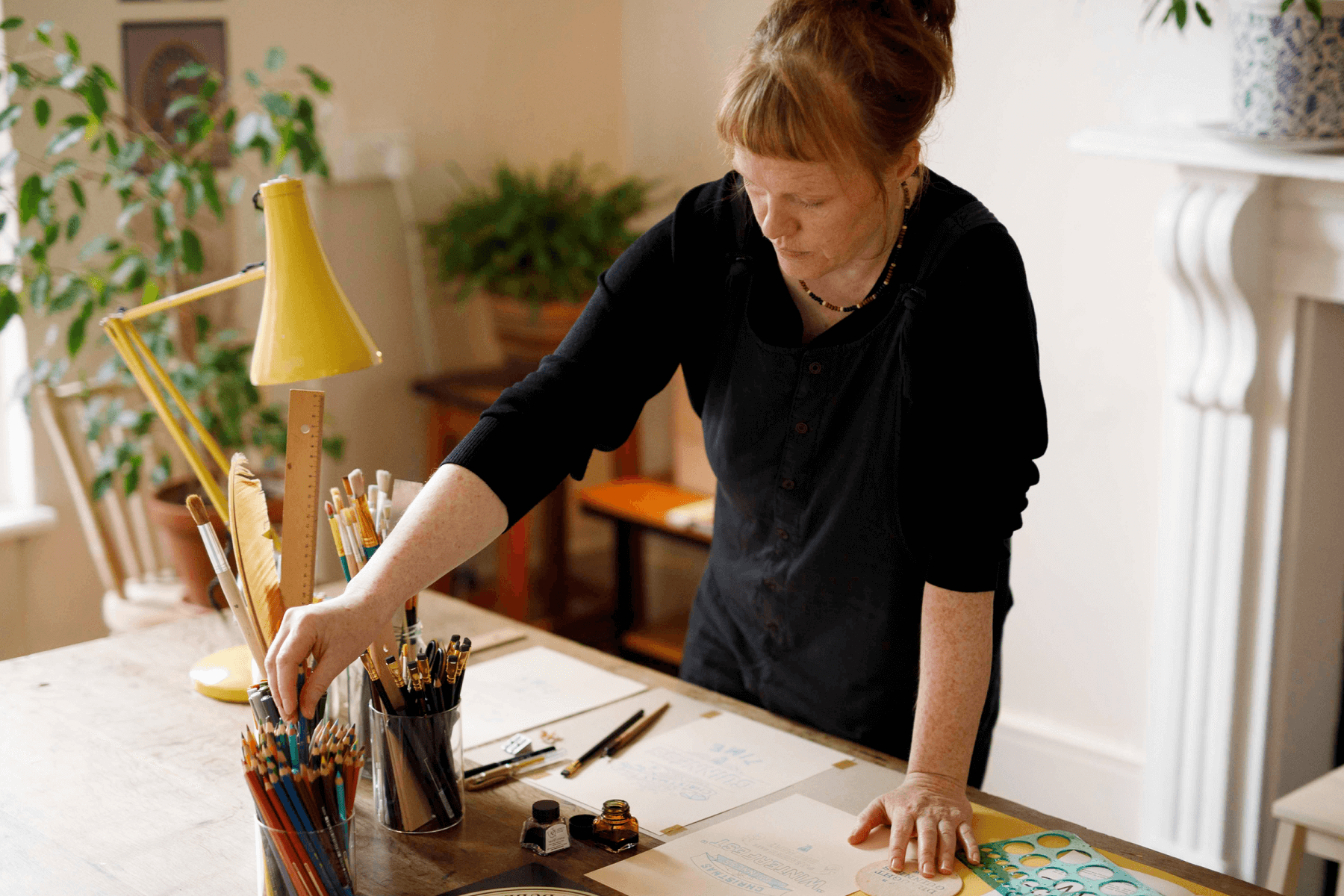 Annie Atkins standing in front of a work table splayed with her designs reaching for colored pencils