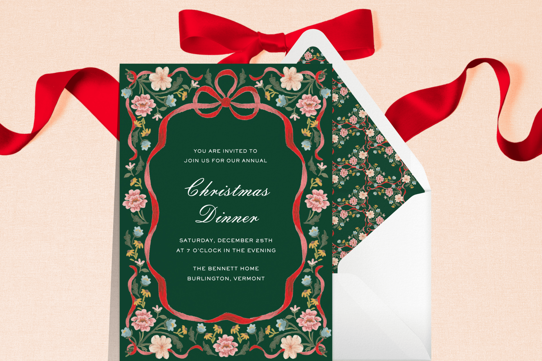 A dark green Christmas invitation featuring a red ribbon and floral border in front of an envelope with a matching floral liner, over a backdrop with a red ribbon.