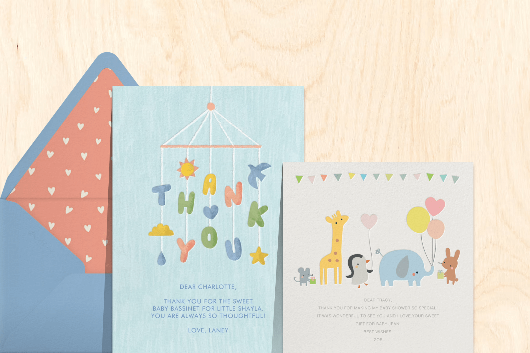 Two baby-themed thank you cards—one with a crib mobile and one with cartoon animals carrying balloons—nrext to a blue envelope with hearts liner and a woodgrain backdrop.