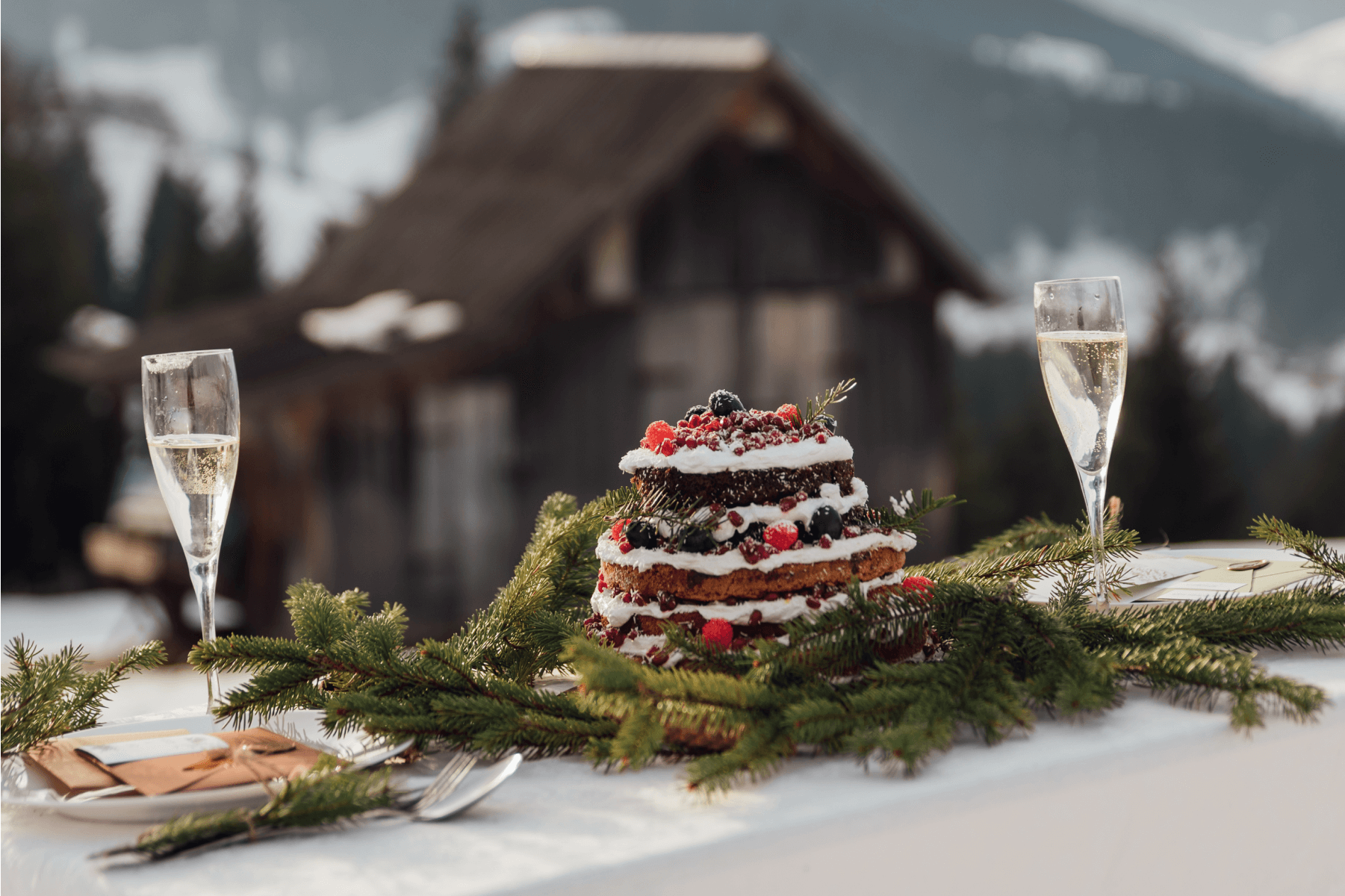 A table set with green branches, Champagne glasses, and a layered cake topped with red berries in front of a cabin. 