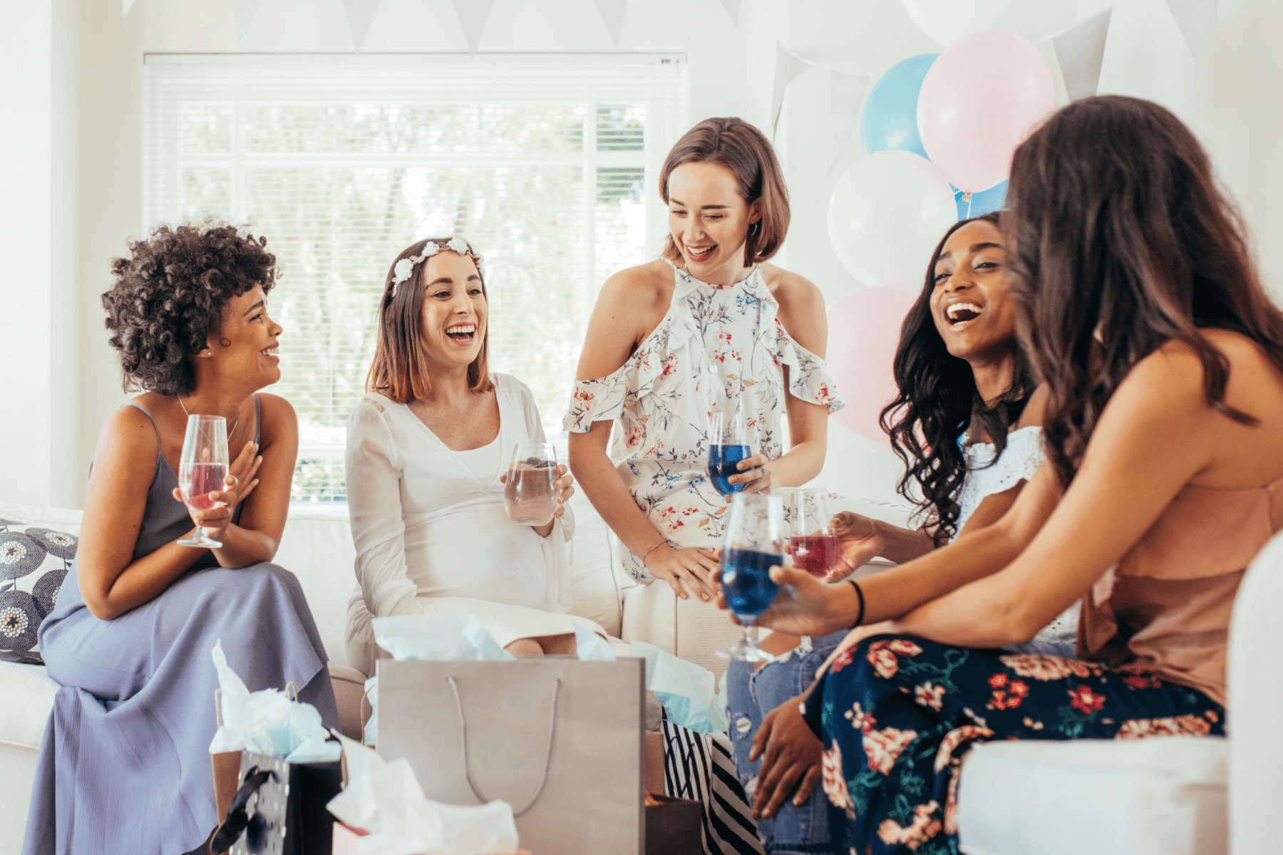 A group of women at a baby shower laughing, drinking wine, and opening presents.