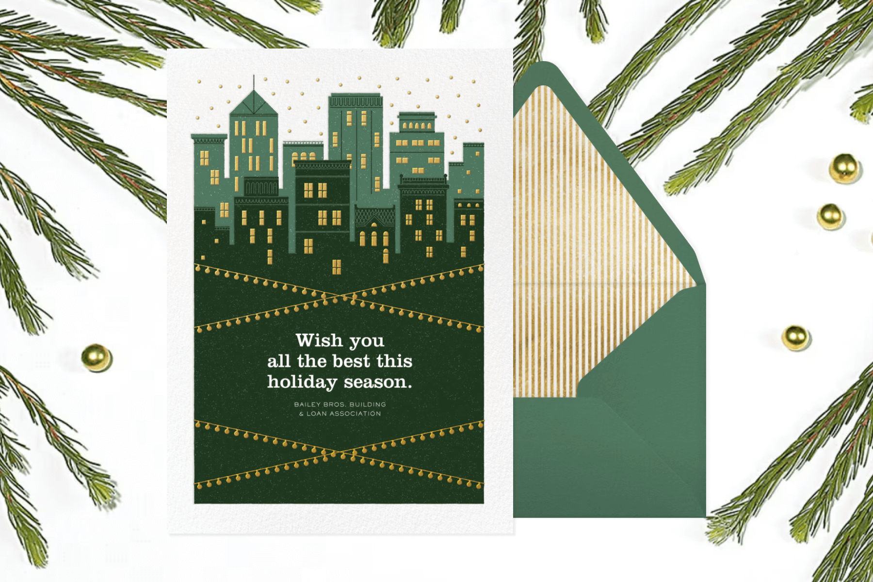 White holiday card with green city skyline and a green envelope on top of a white background with pine needles