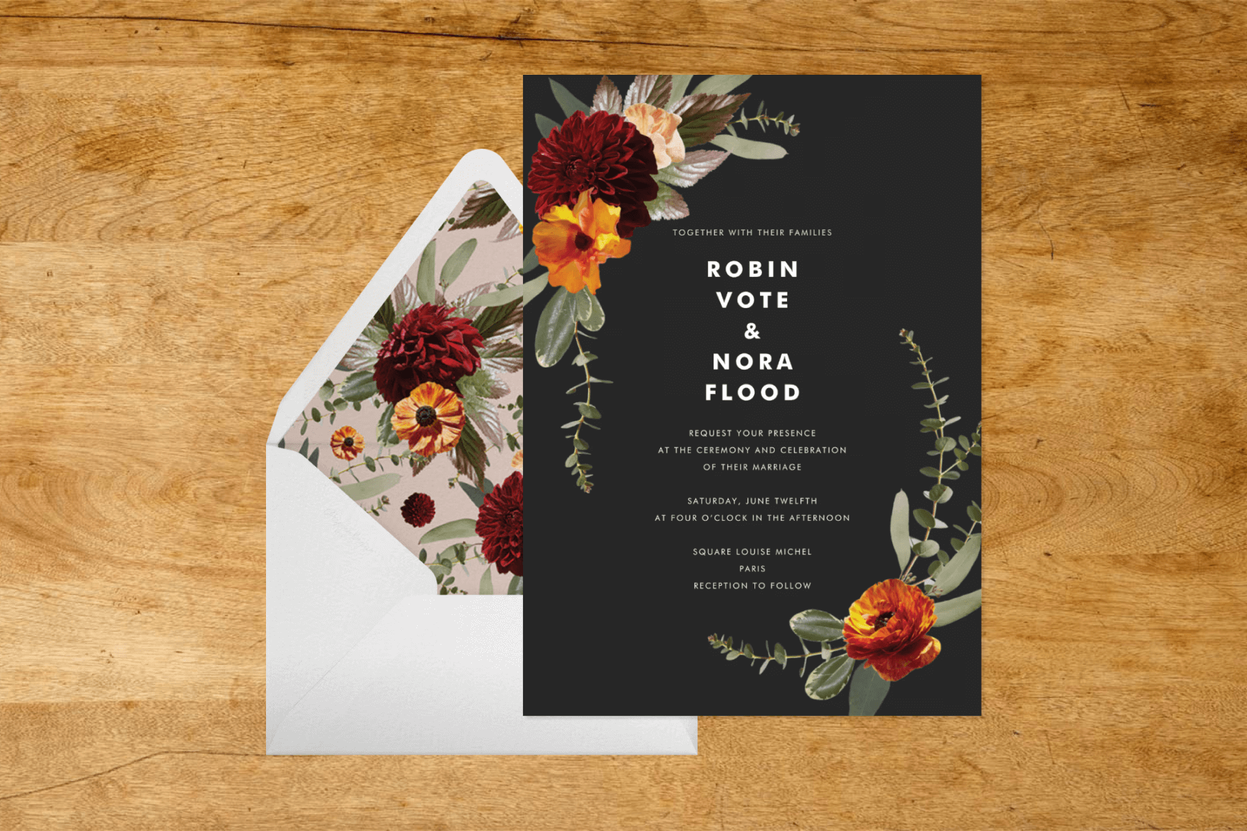 A wedding invitation with white text on a black background and a floral border design, a white envelope with a floral liner, and a wooden background. 