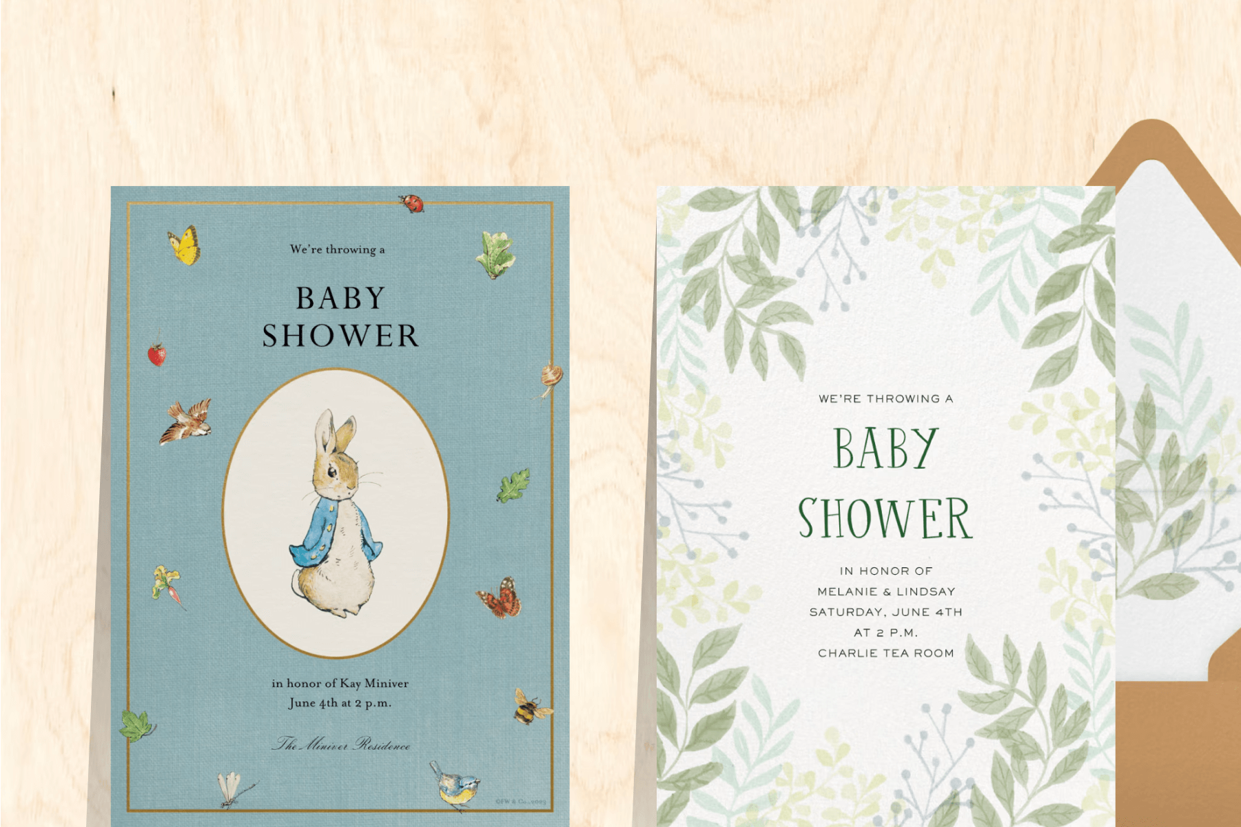 A blue baby shower invitation with Peter Rabbit surrounded by butterflies and birds, and a white baby shower invitation with framing greenery, both over a wooden backdrop.