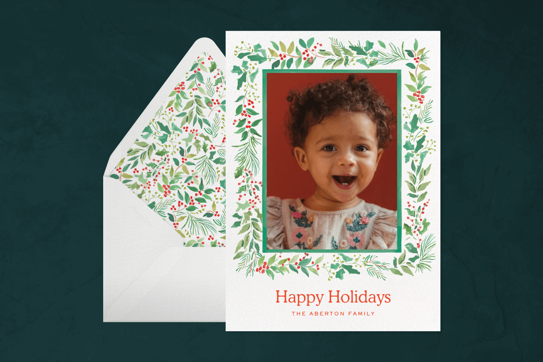 A holiday card featuring a baby photo in a garland frame, in front of an envelope with a matching garland liner.