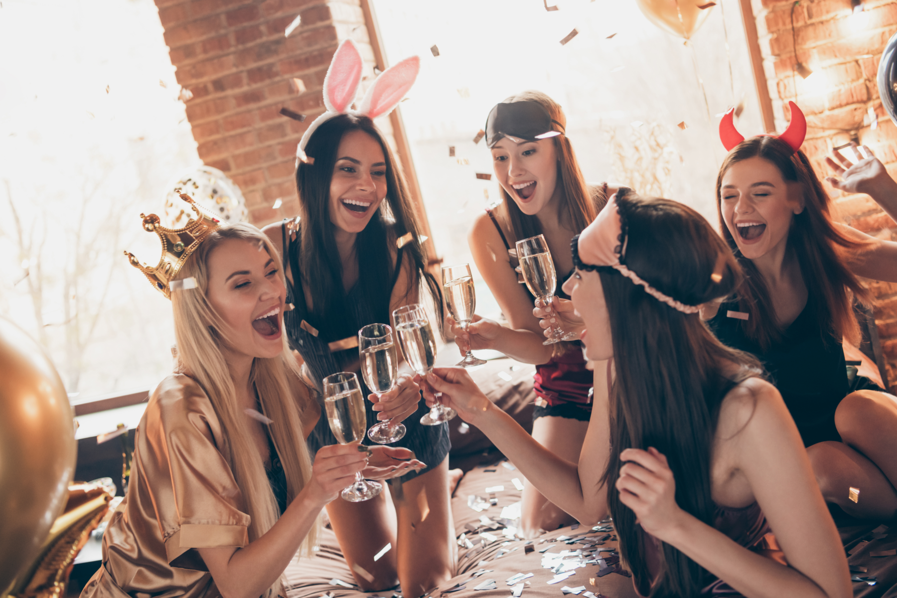 Women toast with Champagne flutes while wearing playful costume headbands like bunny ears and devil horns.