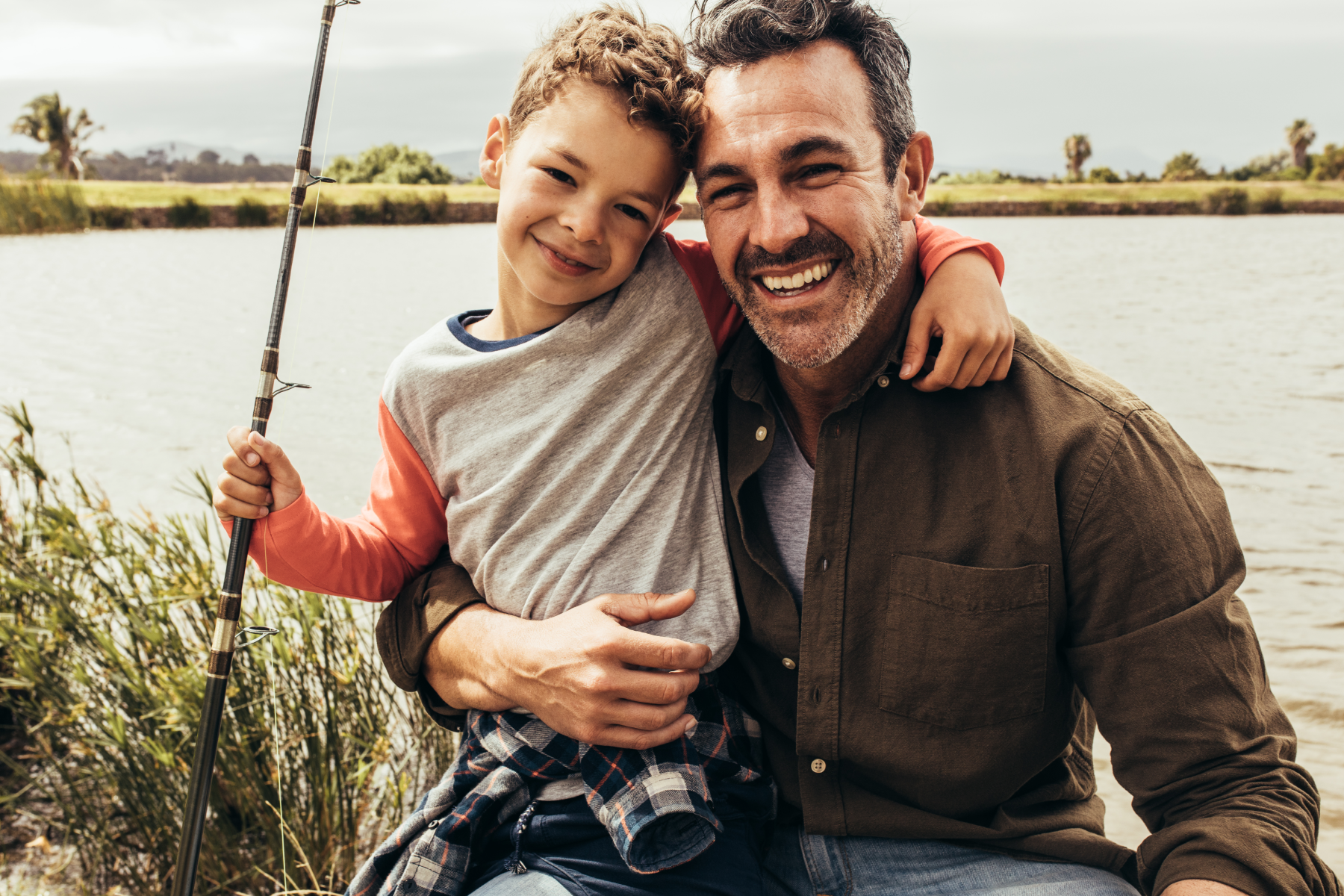 A man and boy are fishing and embracing in front of a lake.